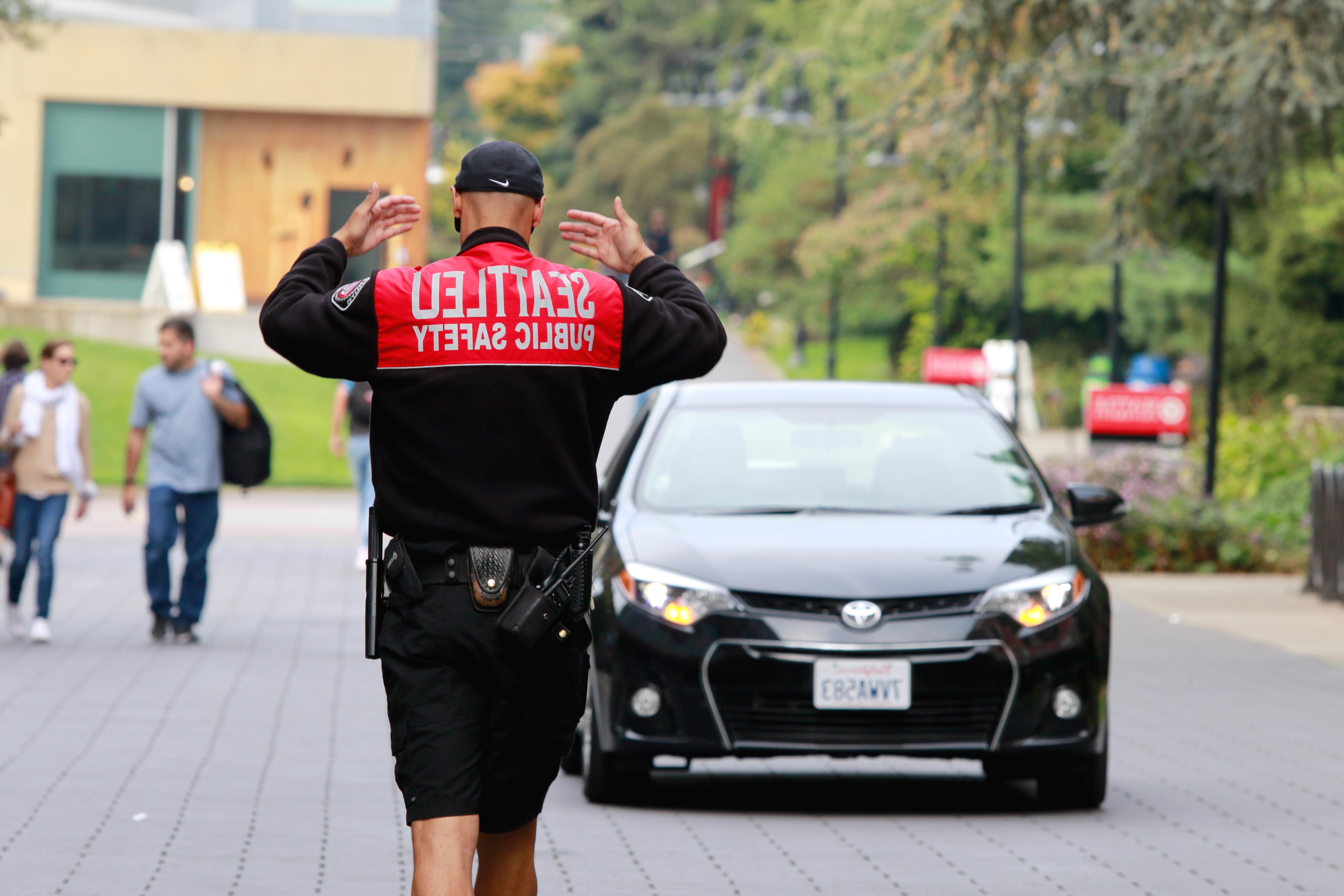 DPS officer directing traffic with both arms up. Back facing camera. A black sedan is driving toward the officer.